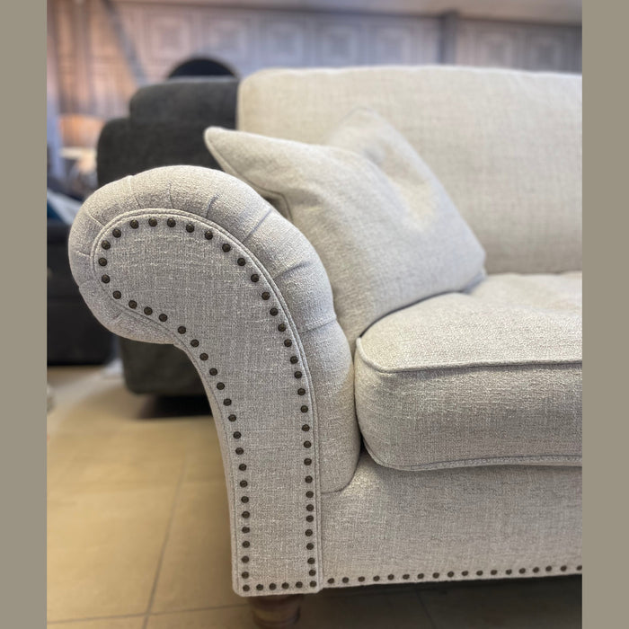 A close-up of the light gray Keaton sofa shows rolled armrests, nailhead trim, wooden legs, and a fibre-filled seat cushion. In the background, a dark sofa is visible.