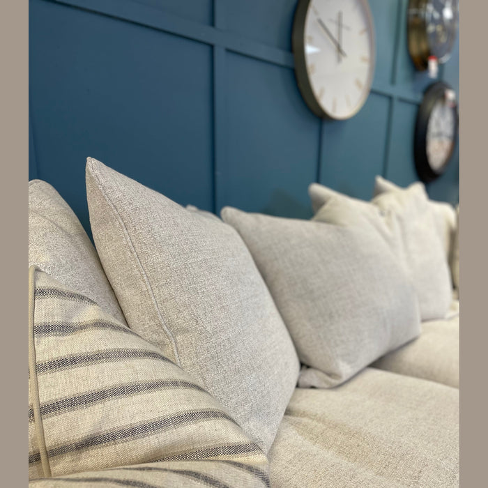 A close-up of the Keaton sofa, upholstered in beige fabric with fibre-filled seats and assorted cushions—including a gray-striped one—set against a dark blue paneled wall topped with round clocks.