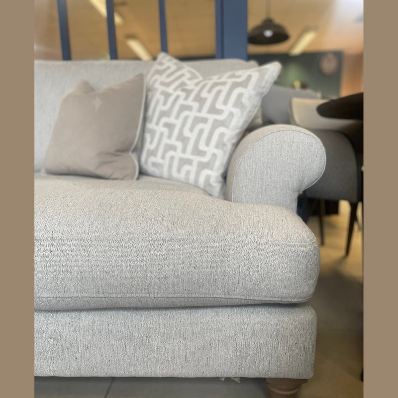 A light gray Monroe Sofa featuring Irish fabric band detailing and two patterned throw pillows, set indoors with a blue partition and tables visible in the background.