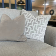 Two decorative pillows sit on a light gray upholstered armchair from the Monroe Sofa Range—one in plain beige, the other light gray with a white geometric pattern. A softly blurred interior appears in the background.