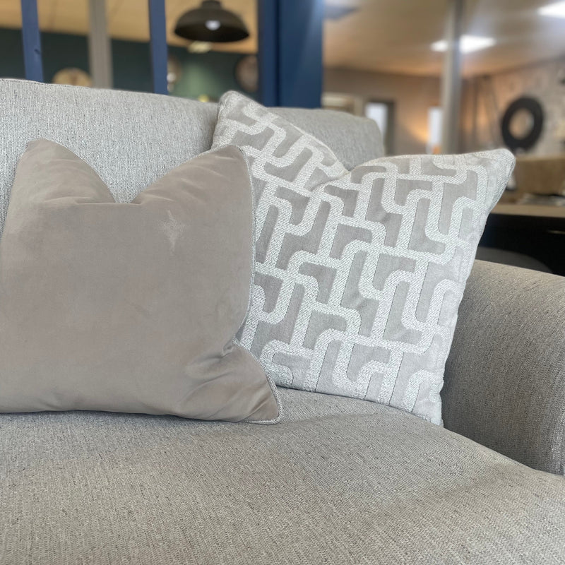 Two decorative pillows sit on a light gray upholstered armchair from the Monroe Sofa Range—one in plain beige, the other light gray with a white geometric pattern. A softly blurred interior appears in the background.
