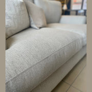 Close-up of a light gray Monroe Sofa Range piece with textured Aqua Clean fabric, cushion detail, and tiled floor underneath. Blurred furnishings and framed decor in the background emphasize the fabric’s durability.