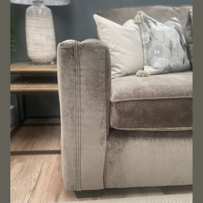 A close-up of the Ariya corner sofa in plush, light gray velvet-like fabric offers maximum comfort. Decorative pillows add style. Beside it, a wooden side table with a woven lamp creates a cozy, modern living room vibe.