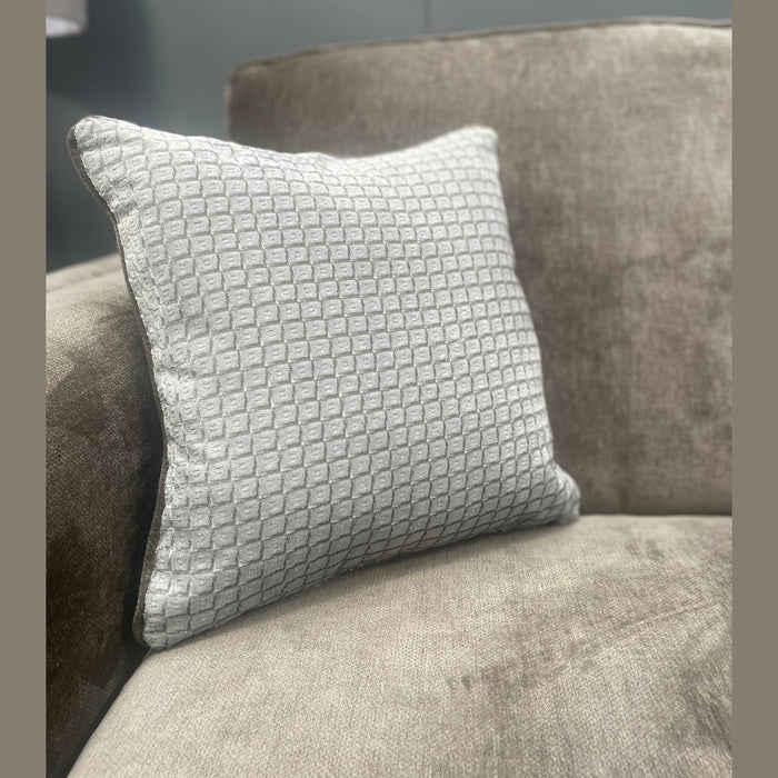 A light gray textured cushion sits on the arm of an Ariya Corner Sofa in soft beige, providing comfort in the living room, with a blurred gray wall and window behind.