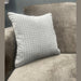 A light gray textured cushion sits on the arm of an Ariya Corner Sofa in soft beige, providing comfort in the living room, with a blurred gray wall and window behind.