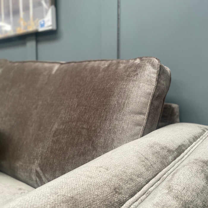 A close-up of the Ariya sofa highlights its plush grey upholstery and a dark pillow, set against a matte blue-gray wall. The scene, ideal for cozy living rooms, is accented by framed artwork in the top left corner.