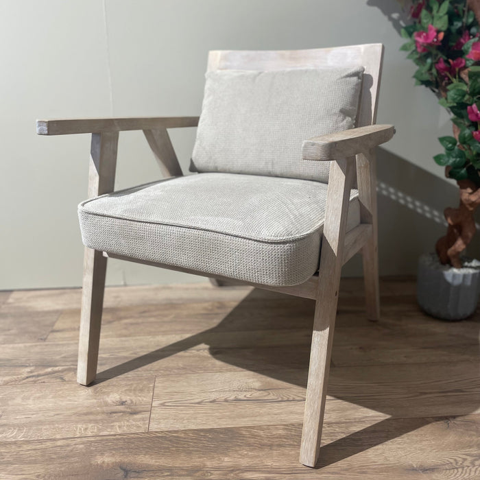 The Willow Chair, featuring beige upholstery, sits on a wooden floor beside a pink-flowered plant and a light-colored wall.