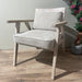 The Willow Chair, featuring beige upholstery, sits on a wooden floor beside a pink-flowered plant and a light-colored wall.