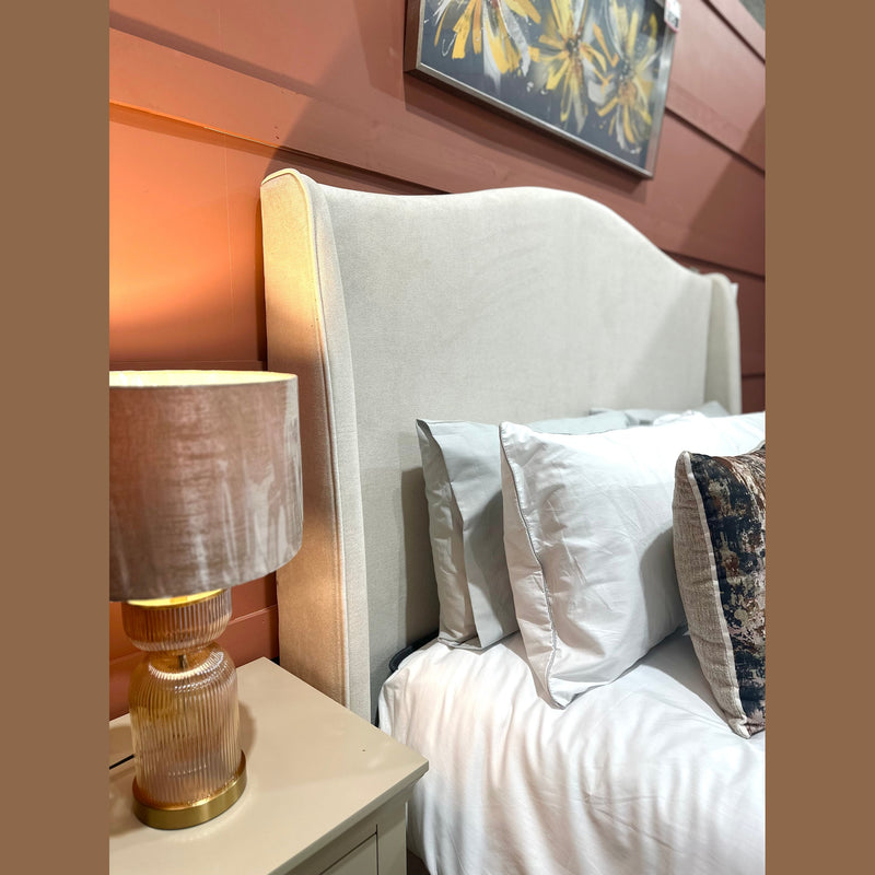 The Venice Bedframe 5ft features elegant cream upholstery and a curved headboard, styled with white pillows and a patterned cushion. Positioned next to a nightstand with a textured lamp, this modern luxury bed is completed by floral framed artwork hanging on the wall above.