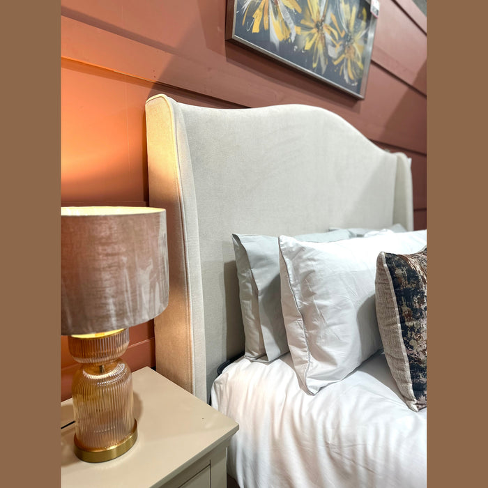 A cozy bedroom with the Venice Bedframe in cream upholstery and a curved headboard, white pillows, a patterned cushion, a bedside table, and a gold-accented lamp, set against rust-hued paneled walls topped with floral artwork.