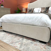 The Venice Bedframe 5ft, with its cream upholstery and gracefully curved headboard, takes center stage in the cozy bedroom. It is dressed in crisp white bedding and decorative pillows, complemented by a patterned rug. A dresser, lamp, and a green plant complete the inviting backdrop.