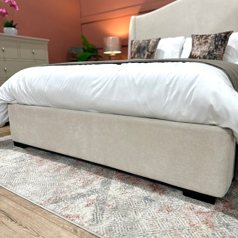 The Venice Bedframe 5ft, with its cream upholstery and gracefully curved headboard, takes center stage in the cozy bedroom. It is dressed in crisp white bedding and decorative pillows, complemented by a patterned rug. A dresser, lamp, and a green plant complete the inviting backdrop.