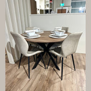 The Elson Round Table with modern black X legs is paired with 4 beige-cushioned Lyra Chairs on a light wooden floor. Each setting includes a white plate, bowl, cup, and utensils atop woven placemats.