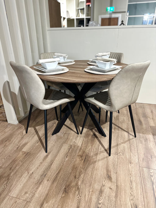 The Elson Round Table with modern black X legs is paired with 4 beige-cushioned Lyra Chairs on a light wooden floor. Each setting includes a white plate, bowl, cup, and utensils atop woven placemats.