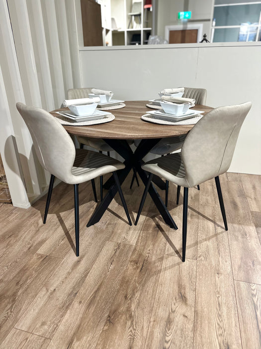 The Elson Round Table with modern black X legs is paired with 4 beige-cushioned Lyra Chairs on a light wooden floor. Each setting includes a white plate, bowl, cup, and utensils atop woven placemats.