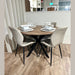 The Elson Round Table with modern black X legs is paired with 4 beige-cushioned Lyra Chairs on a light wooden floor. Each setting includes a white plate, bowl, cup, and utensils atop woven placemats.