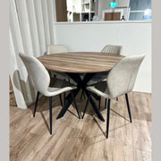 The Elson Round Table with black X legs is paired with four Lyra light beige padded chairs, all set on a light wood floor in a modern, minimal space.