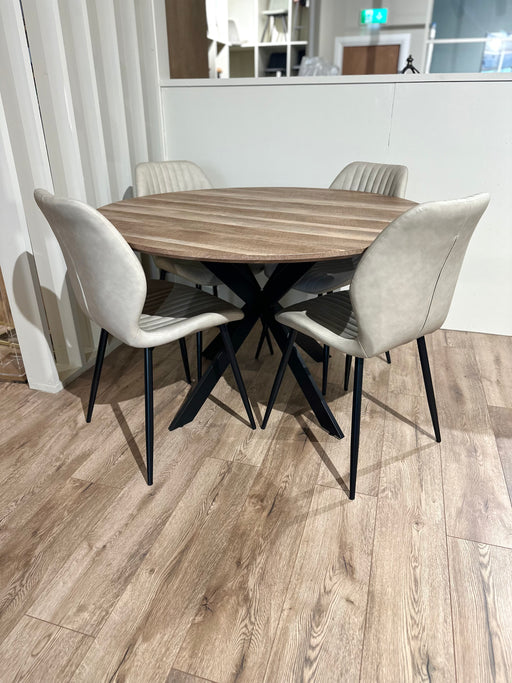 The Elson Round Table with black X legs is paired with four Lyra light beige padded chairs, all set on a light wood floor in a modern, minimal space.
