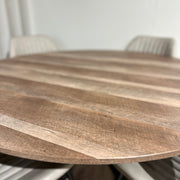 The Elson Round Table with black X legs and a textured wood surface is paired with four light-colored, padded Lyra Chairs, creating a modern, minimalistic dining space.