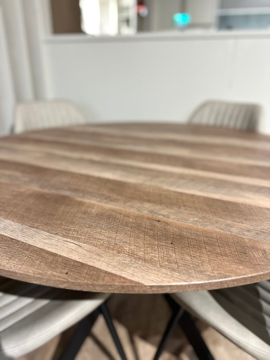 The Elson Round Table with black X legs and a textured wood surface is paired with four light-colored, padded Lyra Chairs, creating a modern, minimalistic dining space.