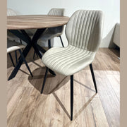 The Elson Round Table with 4 Lyra Chairs features modern dining chairs with light beige, vertically-stitched upholstery and black metal legs, arranged around a round table on a light brown wooden floor.