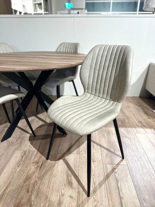 The Elson Round Table with 4 Lyra Chairs features modern dining chairs with light beige, vertically-stitched upholstery and black metal legs, arranged around a round table on a light brown wooden floor.
