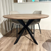 The Elson Round Table with four Lyra Chairs features black X legs and light gray padded seats, creating a modern, minimalistic dining setup on a light wood floor.