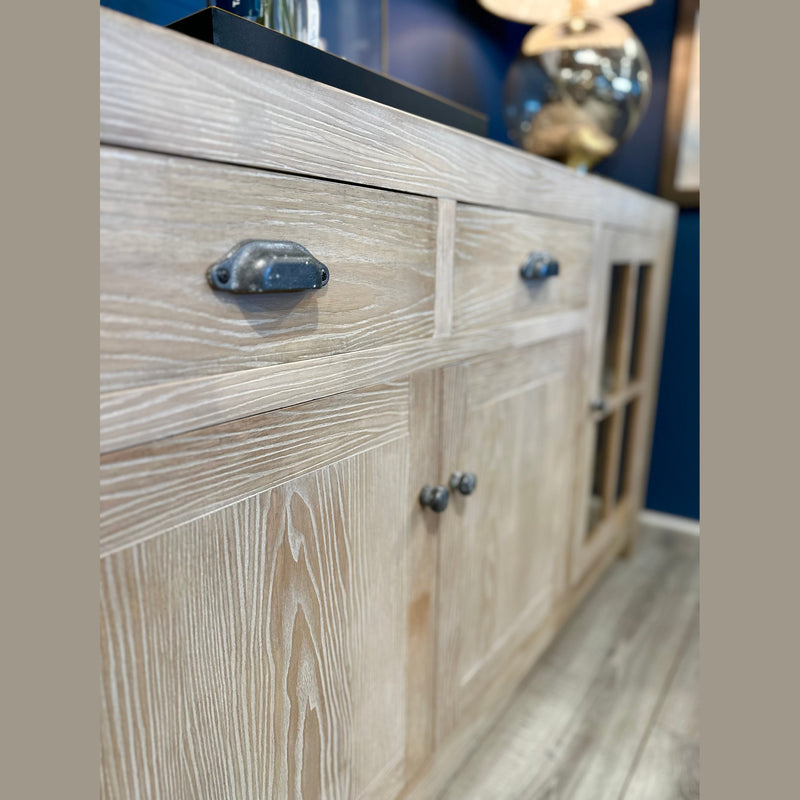 A close-up of the Le Manoir Sideboard highlights its visible grain patterns, drawers with metal handles, and glass-paneled cabinet doors, set against a blue wall on a wooden floor.