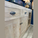 A close-up of the Le Manoir Sideboard highlights its visible grain patterns, drawers with metal handles, and glass-paneled cabinet doors, set against a blue wall on a wooden floor.