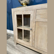 A close-up of the Le Manoir Sideboard, featuring light wood, a glass-paneled door with a metal knob, and visible interior shelves, standing on a wood floor against a blue wall.