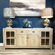 The Le Manoir Sideboard with glass doors displays two metallic table lamps with beige shades, a small flower vase, and candles. A landscape painting is mounted on the dark blue wall behind the arrangement.