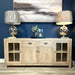 The Le Manoir Sideboard with glass doors displays two metallic table lamps with beige shades, a small flower vase, and candles. A landscape painting is mounted on the dark blue wall behind the arrangement.