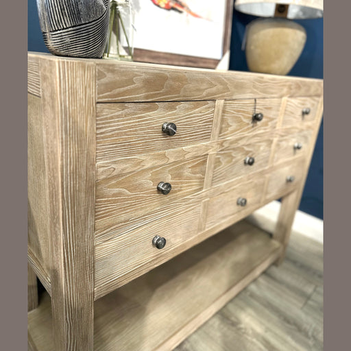 The Le Manoir 9 Drawer Cabinet features a light wood grain finish with dark round knobs. It stands on a wooden floor and is topped with a lamp and decor, while framed art hangs on the navy blue wall behind for a modern look.