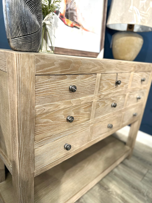 The Le Manoir 9 Drawer Cabinet features a light wood grain finish with dark round knobs. It stands on a wooden floor and is topped with a lamp and decor, while framed art hangs on the navy blue wall behind for a modern look.