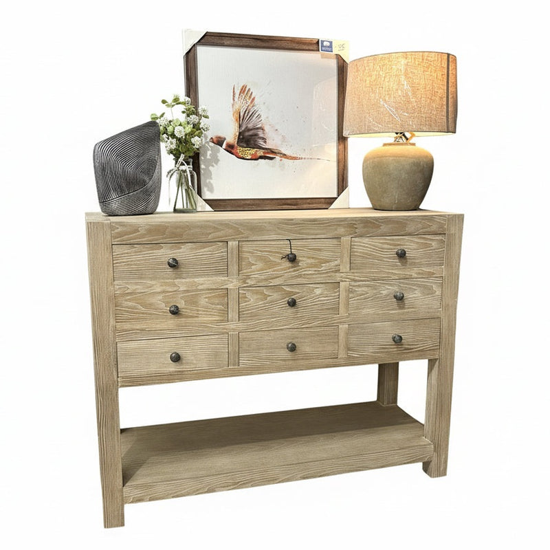 The Le Manoir 9 Drawer Cabinet sits on a wood floor against a blue wall, topped with a beige lamp, framed bird art, metal décor, and a white flower vase—an elegant modern storage piece showcasing its rich wood grain.