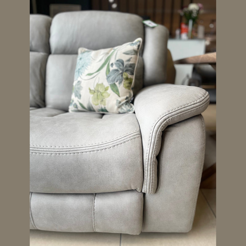 A light gray Reno Recliner Sofa (3 seater) with washable fabric features a floral cushion. It sits on a tiled floor, with a table of flowers softly blurred in the background.