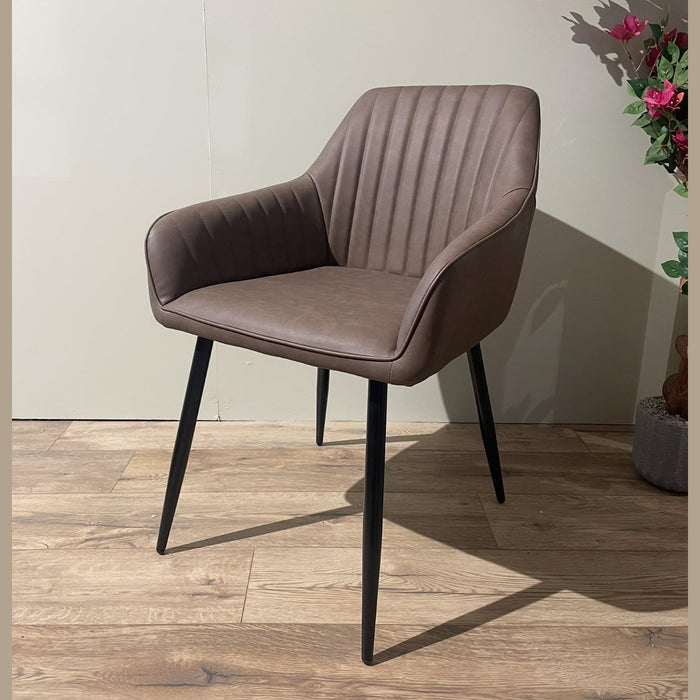 The Enzo Dining Chair features brown leatherette upholstery, vertical-stitched backrest, cushioned seat, black metal legs, and armrests. It is placed on a wooden floor beside a plant with pink flowers.