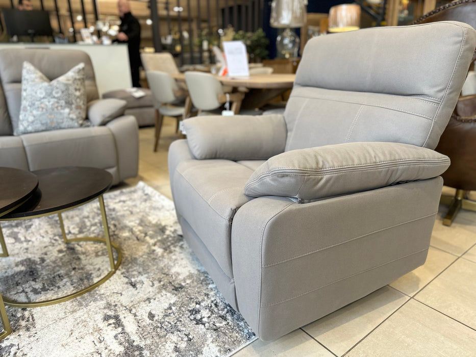 The Jude Recliner, featuring a modern design and light gray washable microfiber, is displayed on a patterned area rug in a showroom with other chairs, tables, and décor.