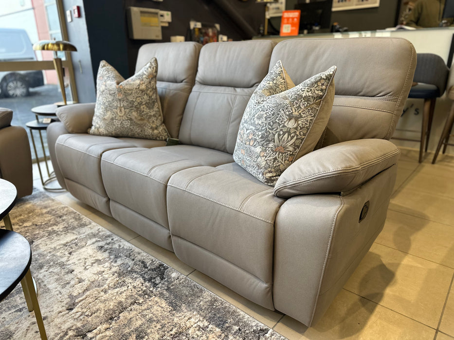 The Jude Recliner is a modern light gray three-seater sofa with washable microfiber fabric and two floral-patterned throw pillows, displayed on a tiled floor beside a window and patterned rug in a contemporary living room.