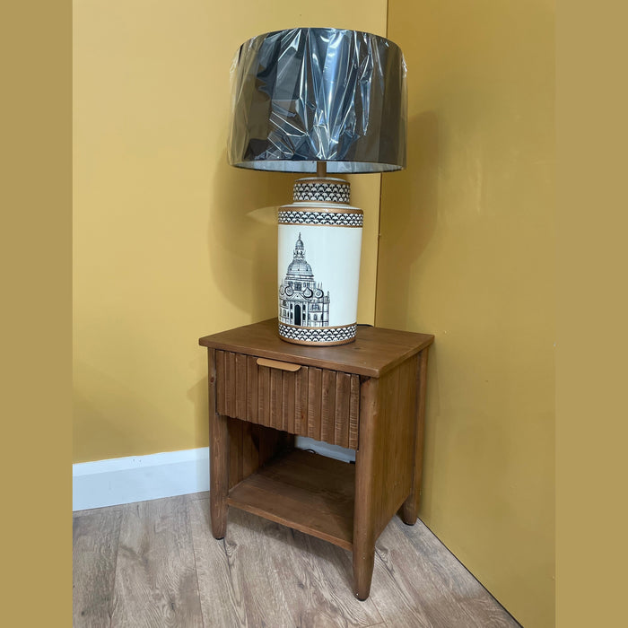 The Cairo Bedside Locker stands against a yellow wall, topped with a white ceramic lamp featuring a black architectural design and a plastic-wrapped shade. A light wood floor completes the scene.