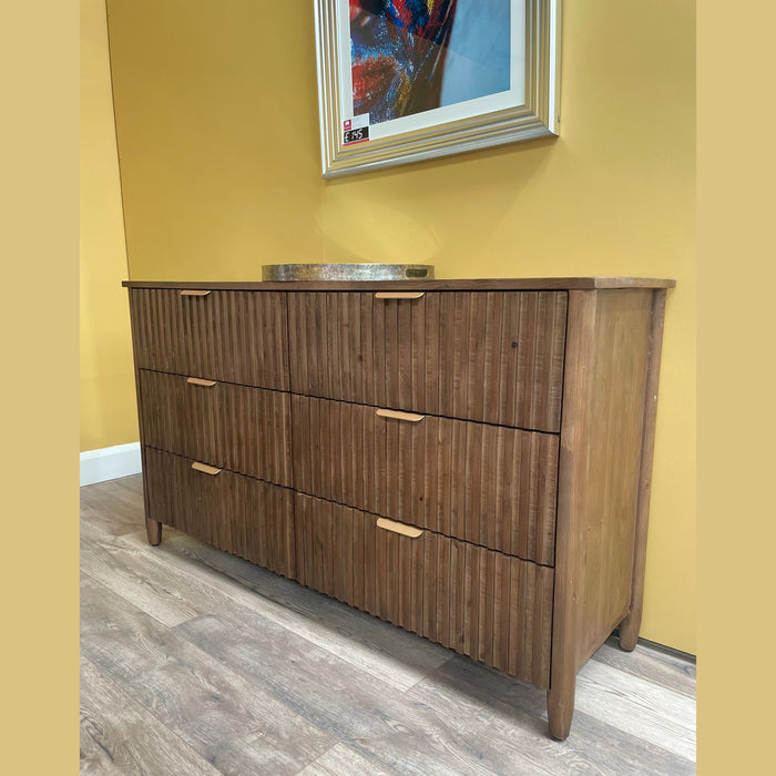 The Cairo Wide Chest, a sustainable wooden dresser with six drawers and vertical grooves, stands against a yellow wall. A round tray sits on top with a framed picture above, all on a light wood floor.