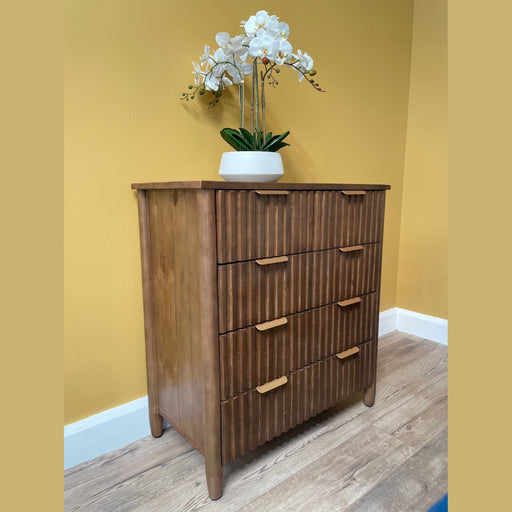 The Cairo Tall Chest from a rustic bedroom collection, featuring four vertical-grooved drawers, stands against a yellow wall on a light wood floor with a white pot of blooming orchids placed on top.