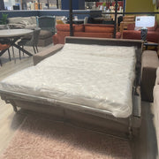 The Sqaured Sofabed, a modern brown sofa bed with a pocket sprung mattress, is displayed extended and wrapped in plastic on a light pink rug in a furniture store among various sofas, chairs, and tables.