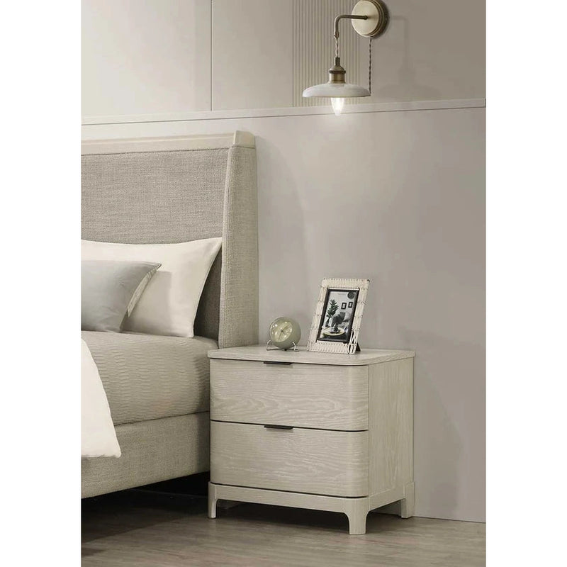 A beige bedside locker placed next to a bed, featuring two drawers and a handle, with a wall lamp above it.