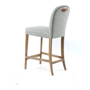 The Jenny Counter Stool features a cream fabric upholstered seat and backrest, wooden legs, metal stud trim, and a small brown leather handle on the backrest for extra charm.