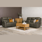A modern living room with two dark gray leather sofas, including the Russo Electric Recliner - Leather, decorated with throw pillows, a wooden coffee table with books and a candle, a wooden cabinet with vases and greenery, and a light rug on wood flooring.