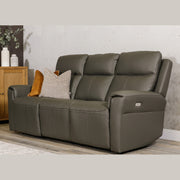 A Russo Electric Recliner - Leather in dark gray with built-in USB charging and two neutral pillows rests on a wooden floor by a cabinet with vases and greenery, set against a light textured wall.