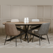 The Sadie Oval Dining Table with a wooden top and black metal legs is paired with four neutral upholstered chairs. Three white ceramic vases sit at the center, complemented by beige paneled walls and wood flooring.