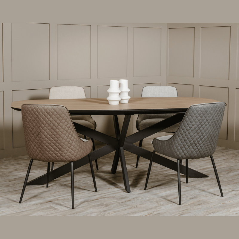 The Sadie Oval Dining Table with a wooden top and black metal legs is paired with four neutral upholstered chairs. Three white ceramic vases sit at the center, complemented by beige paneled walls and wood flooring.