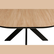 A close-up of the Sadie Oval Dining Table shows its light brown, smooth wooden surface and black metal X-shaped legs, photographed from above with a focus on one side.